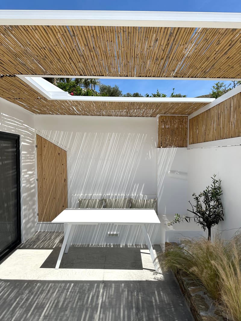 The courtyard under the reed pergola, open to the sky, with the table, cushioned bench and glazed door