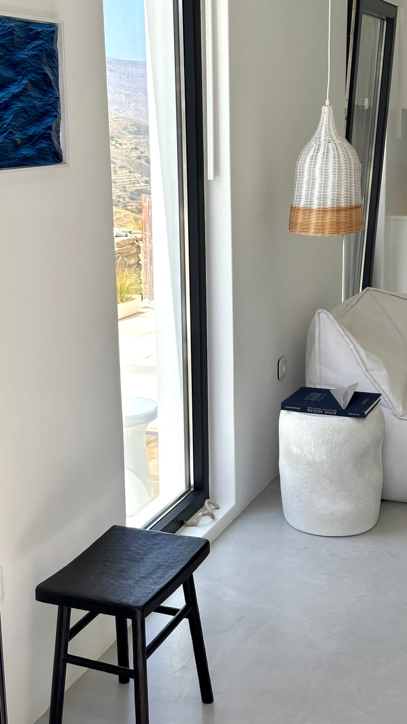 Ψηλό παράθυρο με θέα στους λόφους, μαύρο σκαμπό και ψάθινο κρεμαστό φωτιστικό